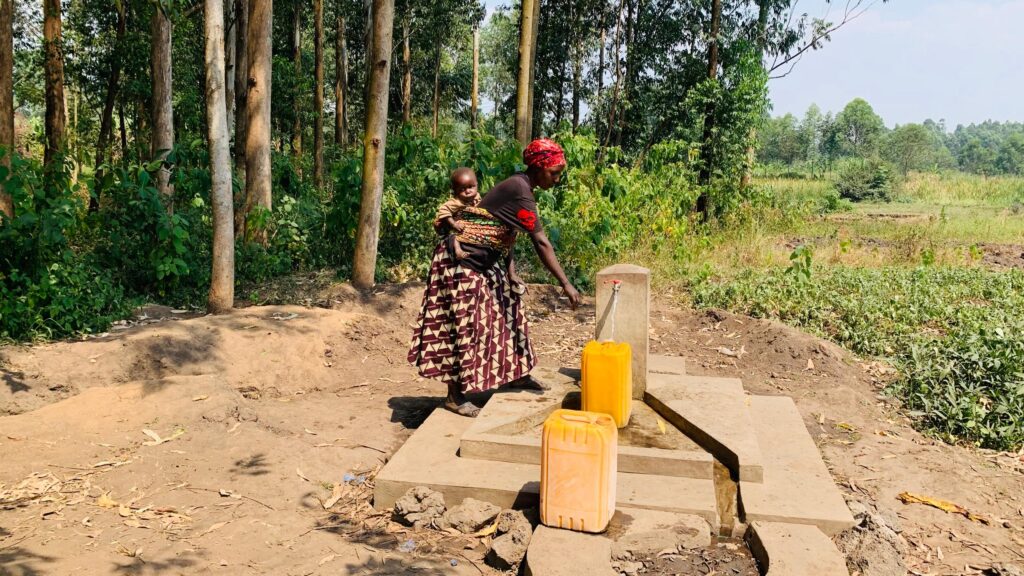 A woman carries a child on her back while fetching water from a rural pump. Two yellow jerry cans are placed nearby. The setting is a forested area.