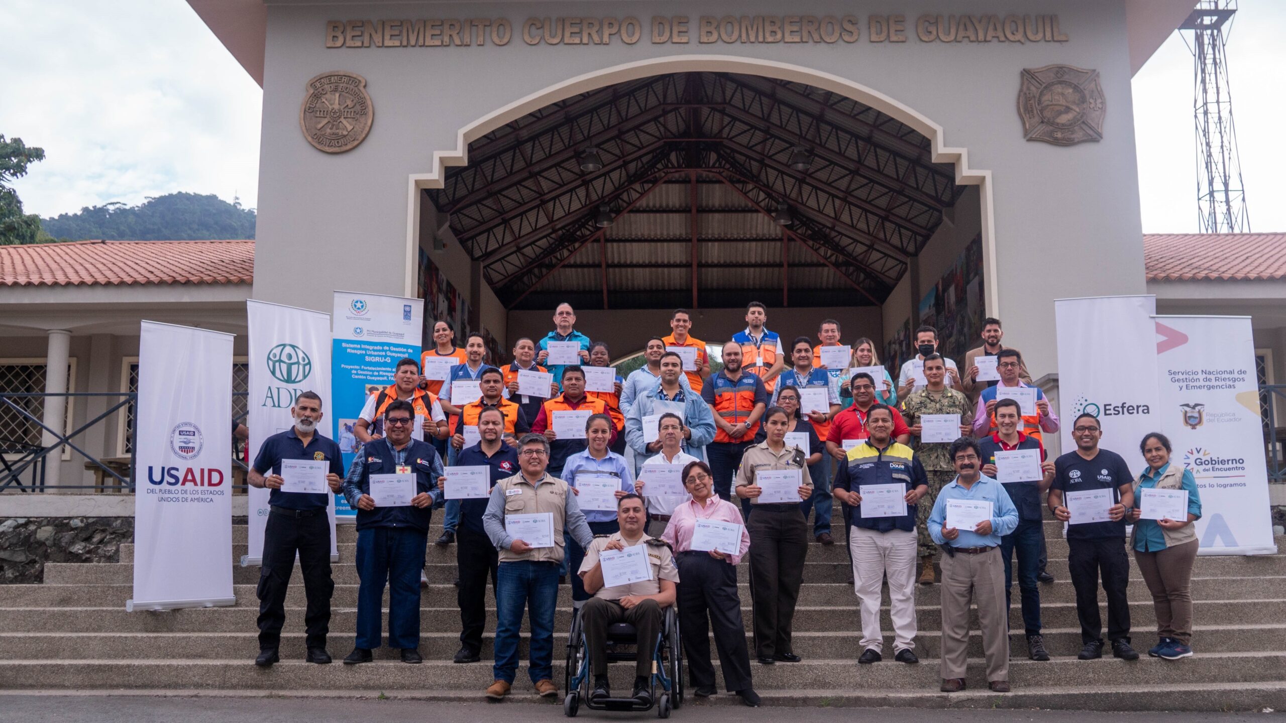 Un grupo de 37 personas posa en las escaleras frente a un edificio, sosteniendo certificados en sus manos. Una persona está en silla de ruedas. Pueden verse rollups publicitarios de USAID, ADRA y Esfera. En la fachada del edificio pueden leerse las palabras “Benemérito Cuerpo de Bomberos de Guayaquil”